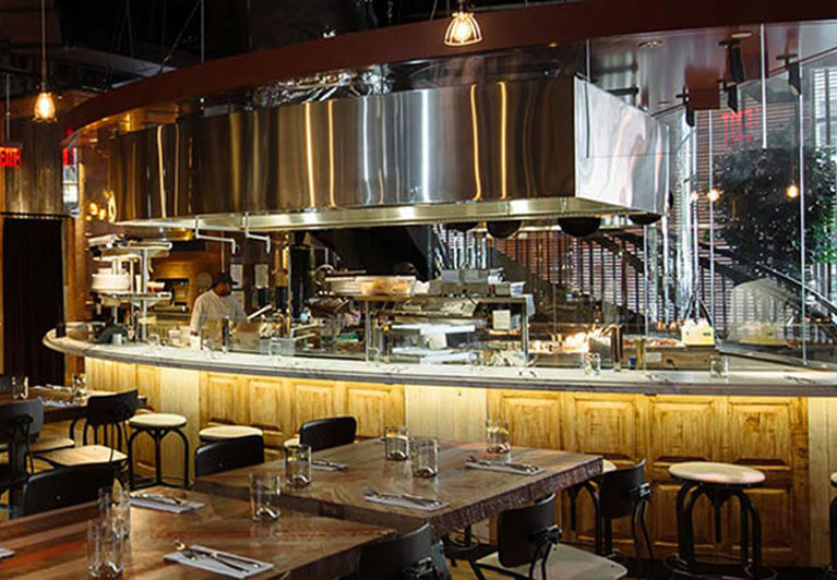 Restaurant vide avec des tables en bois et une personne travaillant derrière une cuisine ouverte en forme de courbe