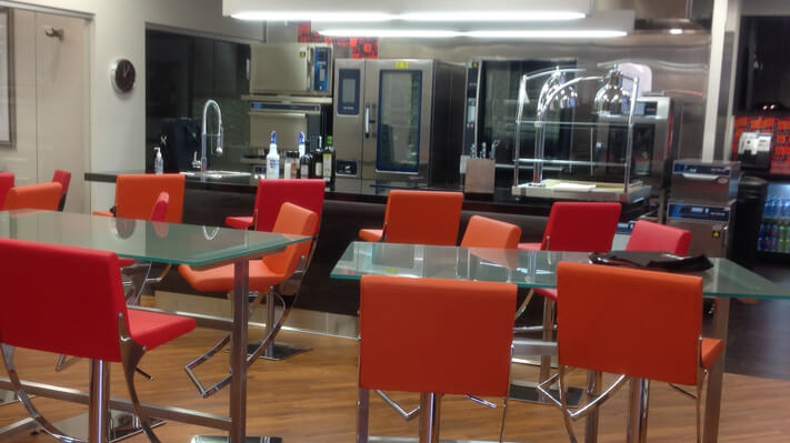 Kitchen with a counter, sink, commerical appliances amd deep orange bar stools arranged around two glass tables